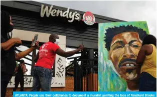  ?? — AFP ?? ATLANTA: People use their cellphones to document a muralist painting the face of Rayshard Brooks outside a burned Wendy’s restaurant on Sunday.