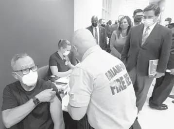  ?? WILFREDO LEE/AP ?? Florida Gov. Ron DeSantis, right rear, watches as Carlos Dennis, left, 65, rolls up his sleeve so that Miami-Dade County Fire Rescue paramedic, Capt. Javier Crespo, can administer a COVID-19 vaccine shot Monday at Jackson Memorial Hospital in Miami.