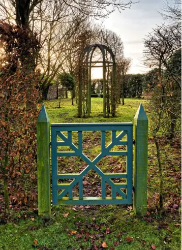  ??  ?? A cluster of red-twigged lime, Tilia ‘Rubra’, catches the January sunlight. A decorative wooden gate, blending with the garden colours, mirrors the geometric style of the layout. Beyond is a wooden gazebo with a vista through to another obelisk. The bare hedges are of hornbeam, Carpinus betulus.