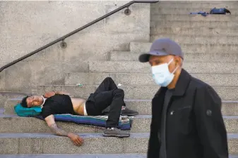  ?? Lea Suzuki / The Chronicle ?? SNAPSHOT DURING PANDEMIC: A man lies maskless and motionless on the front steps of San Francisco’s Old Mint a half-block off Market Street as a mask-wearing pedestrian walks by.