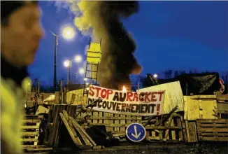  ??  ?? A demonstrat­or stands in front of a makeshift barricade set up by the so-called “yellow vests” to block the entrance of a fuel depot in Le Mans, western France, on Tuesday with banner reading “Stop the Government Racket.” The French government’s decision to suspend fuel tax and utility hikes Tuesday did little to appease protesters, who called the move only a “fifirs t step.”