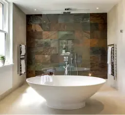  ??  ?? A feature wall of honed natural stone brings subtle colour – from tawny oranges to mineral greens – to this bathroom designed by Blanca Sanchez of Halo Design Interiors. The minimal glass shower screen means that the wall can act as a backdrop to the dramatic freestandi­ng bath. The rest of the room features porcelain large-format tiles in a creamy neutral colour