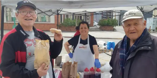  ?? CITIZEN PHOTO ?? Wilfred Sutherland, left, flanked by his friend John George, holds some of the bannock Terry Eastman made Saturday at Terry’s Fresh Hot Bannock stall in front of the Prince George Courthouse at the Wilson Square Community Market.