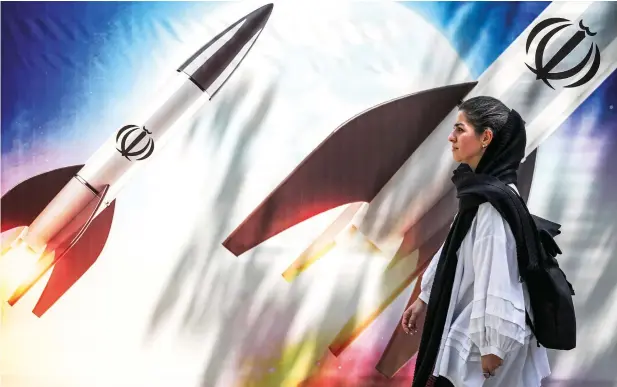  ?? — AFP ?? A woman walks past a banner depicting launching missiles bearing the emblem of the Islamic Republic of Iran in central Tehran on Monday.