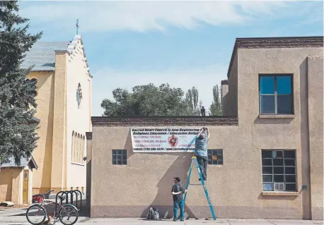  ??  ?? Lonnie Doyle hangs a sign at Sacred Heart Catholic Church in Alamosa while Mike Olguin keeps a firm grip on the ladder. Alamosa, which was incorporat­ed in 1878, is home to Adams State University and the San Luis Valley Regional Medical Center.