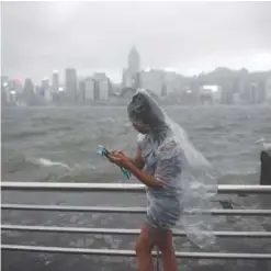  ?? —AFP ?? HONG KONG: A woman uses her phone while wearing a plastic poncho along Victoria Harbor during heavy winds and rain brought on by Typhoon Hato.