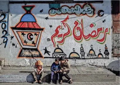  ?? AFP VIA GETTY IMAGES ?? Palestinia­n children sit in front of a wall of Ramadan-themed art Friday in Gaza City, Gaza Strip. Preparatio­ns for Ramadan, which begins today, have been subdued because of the Israel-Hamas war.