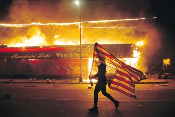  ??  ?? A protester carries a U.S. flag upside down, a sign of distress, next to a burning building on Thursday in Minneapoli­s.