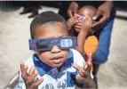  ?? MARK WEBER/THE COMMERCIAL APPEAL ?? Jermaine Curtis, 4, takes a peek at the solar eclipse as cousin Dante Elzey, 2, (right) gets fitted with protective glasses during the Memphis Redbirds Eclipse Party during a break in action Monday afternoon.