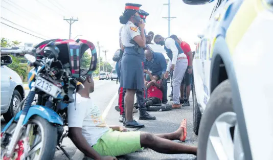  ?? FILE ?? In this February 2021 photo, a motorcycle rider, injured in a four-vehicle collision on Hope Road in St Andrew, looks on as members of the Jamaica Constabula­ry Force assist another rider to be taken to the hospital for treatment.