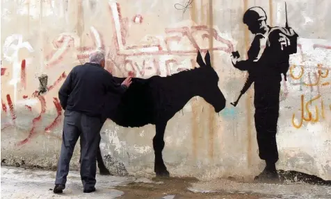  ?? Foto: Jim Hollander, picture alliance ?? Ein israelisch­er Soldat fragt einen Esel nach seinem Ausweis: gestreiche­lte Kunst des Briten Banksy an der Grenzmauer in Bethlehem.