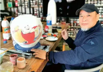  ??  ?? Luo paints a buddha on a pottery piece with piles of bottles of pigments on the table and shelves at the creative center.