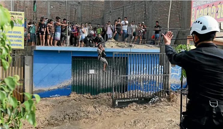  ??  ?? Rescue workers help stranded residents use a zipline to cross a flooded street after the Huayco River burst its banks in Huachipa, Peru. Nearly 70 people have died and more than 100,000 homes have been damaged in flooding caused by unusually heavy El...