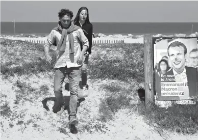  ?? Associated Press ?? A couple runs by defaced election campaign poster for French centrist presidenti­al candidate Emmanuel Macron on Friday on a dune facing the Atlantic Ocean in Bidart, southweste­rn France. Macron will face far-right candidate Marine Le Pen in the...