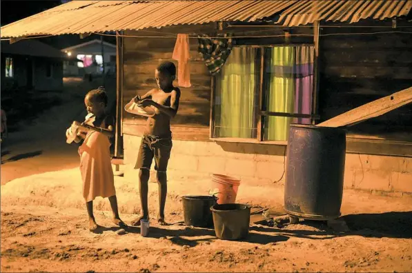  ?? Stephanie Strasburg/Post-Gazette ?? Children clean their shoes for school in the orange glow of a street light in the village of Adjuma Kondre, Suriname. It will take Alcoa years to repair thousands of acres of mined land around the village.