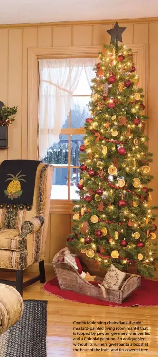  ??  ?? Comfortabl­e wing chairs flank the old mustard-painted living room mantel that is topped by pewter, greenery, cranberrie­s and a Colonial painting. An antique sled without its runners gives Santa a boost at the base of the fruit- and tin-covered tree.