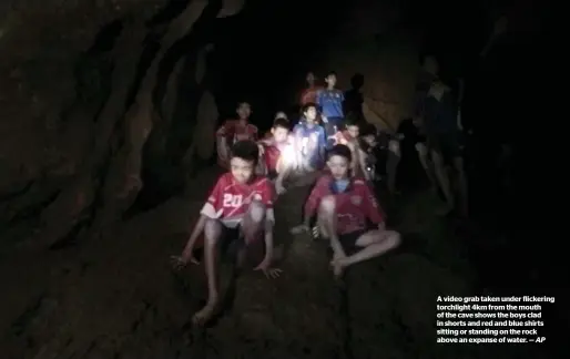  ?? AP ?? A video grab taken under flickering torchlight 4km from the mouth of the cave shows the boys clad in shorts and red and blue shirts sitting or standing on the rock above an expanse of water. —