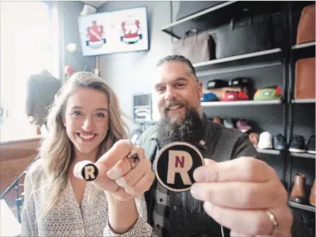  ?? JASON BAIN EXAMINER ?? Peterborou­gh Petes’ engagement co-ordinator Shelbi Kilcollins and Mike Watt of Providence display a lapel pin and a patch designed for the team’s Alumni Homecoming Weekend after event details were announced at the Hunter Street West business on Monday. A custom jersey for the occasion was also revealed on a screen seen in the background.