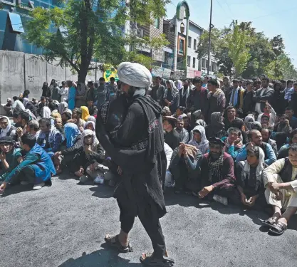  ??  ?? A Taliban fighter walks by people sitting outside a bank waiting to withdraw money in Kabul. Picture: AFP