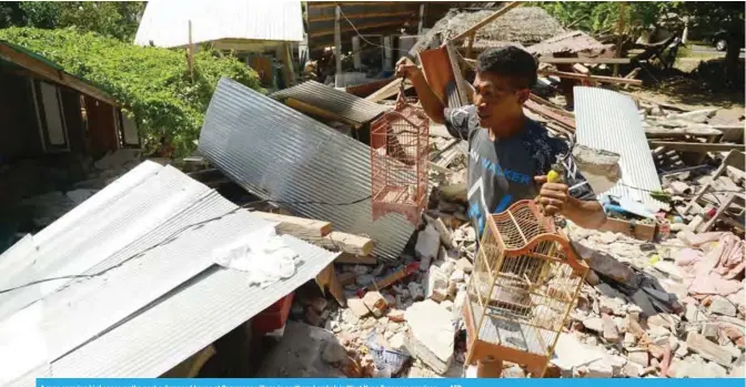  ??  ?? A man carrying bird cages walks past a damaged house at Pemenang village in northern Lombok in West Nusa Tenggara province. — AFP