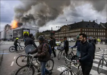  ?? Emil Nicolai Helms/Ritzau Scanpix via AP ?? People ride bicycles as smoke rises from the Old Stock Exchange on Tuesday in Copenhagen, Denmark. One of the city’s oldest structures, known for its elaborate spire of intertwine­d dragon tails, partly collapsed in a large fire.