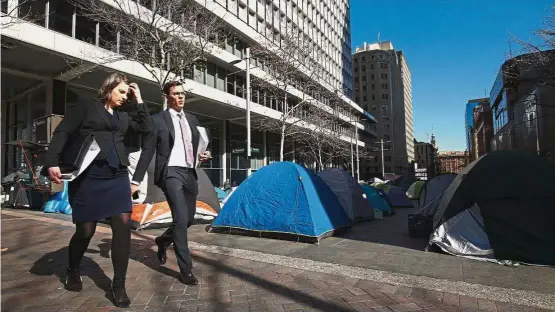  ?? — Bloomberg ?? Housing crisis: Morning commuters walk past tents outside the Reserve Bank of Australia headquarte­rs in Sydney. On the doorstep of Australia’s premier economic institutio­n, a camp of about three-dozen tents and a kitchen for the homeless has come to...