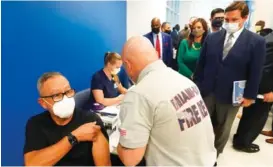  ?? AP PHOTO/WILFREDO LEE ?? Florida Gov. Ron DeSantis, right rear, watches as Carlos Dennis, left, 65, rolls up his sleeve so that Miami-Dade County Fire Rescue paramedic, Capt. Javier Crespo, can administer a COVID-19 vaccine shot at Jackson Memorial Hospital in Miami.
