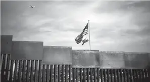  ?? Associated Press ?? ■ U.S. and California state flags fly Monday behind the border wall, seen from Tijuana, Mexico. Tensions have built as nearly 3,000 migrants from a caravan poured into Tijuana in recent days after more than a month on the road—and with many more months likely ahead of them while they seek asylum in the U.S. The federal government estimates the number of migrants could soon swell to 10,000.