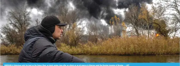  ?? — AFP ?? KHERSON: A fisherman sails his boat on the Dnipro River as black smoke rises after an attack on an oil reserve in Kherson amid the Russian invasion of Ukraine.