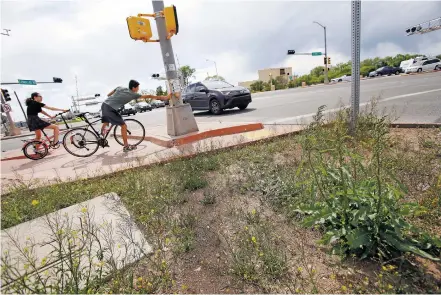  ?? LUIS SÁNCHEZ SATURNO/THE NEW MEXICAN ?? Weeds cover a median at Cerrillos Road and St. Francis Drive on Tuesday as bicyclist Oscar Jaramillo waits to cross with Sarah Deutsch. City spokeswoma­n Lilia Chacon says the ‘extraordin­arily wet winter,’ which was followed by a series of spring showers, compounded the city’s weeds problem.