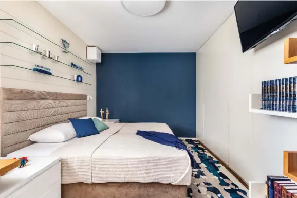  ?? (Orit Arnon) ?? A BLUE accent wall adds a splash of vibrancy to this cozy mamad/ guest room with TV and small shelves. The space is also equipped with a dedicated workspace. Designer: Sandy Brudner | Sandy B Interiors.