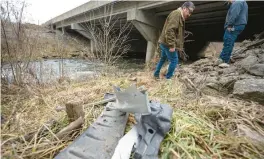  ?? KYLE TELECHAN/POST-TRIBUNE ?? Part of a crushed bumper sits along the shore of Salt Creek in Portage as Nivardo De La Torre and his father-in-law, Mario Garcia, walk the scene where they spotted the wrecked truck that trapped Mishawaka resident Matthew Reum for six days.