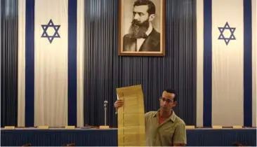  ??  ?? A TOUR guide speaks to visitors next to a mock up of the hall in which Israel’s Declaratio­n of Independen­ce was read. An image of Theodor Herzl hangs over the podium.