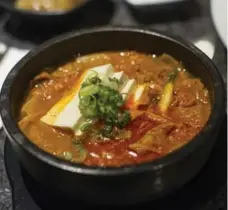  ??  ?? A stew of kimchi and tofu is served bubbling hot in a stone bowl. The thin orange broth contains gleaming rice cakes and shaved pork belly.