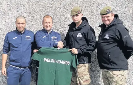  ??  ?? Holyhead Town assistant-manager Darren Harrison and secretary Nick Murphy (furthest left) being presented with warm-up T-shirts from the Welsh Guards at last Saturday’s game.