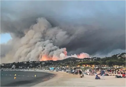  ?? Marion LeFllour, AFP/Getty Images ?? Wildfire evacuees in Bormes-les-Mimosas, France, find refuge on a beach where a massive forest fire provides an ominous backdrop at sunset Wednesday. More than 1,000 firefighte­rs were battling the wildfires.