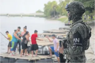  ?? AP ?? A Mexican marine stands guard on the Suchiate River, watching out for migrants crossing from Guatemala to Ciudad Hidalgo, as locals stand on a raft in Mexico on Sunday. Mexico faces heightened pressure from the US to reduce the surge of migrants through its territory.