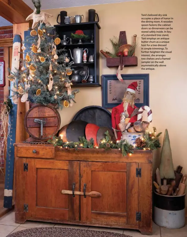  ??  ?? Toni’s beloved dry sink occupies a place of honor in the dining room. A wooden spoon fastens the cabinet doors so dinnerware can be stored safely inside. In lieu of a standard tree stand, Toni employs an antique butter churn as a unique host for a tree dressed in simple trimmings. To further heighten the visual interest, she arranges two shelves and a framed sampler on the wall asymmetric­ally above the antique.