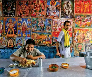  ?? REUTERS ?? A man eats at a roadside stall as a priest walks past a wall with posters of deities at a market area, in Kolkata on Tuesday.