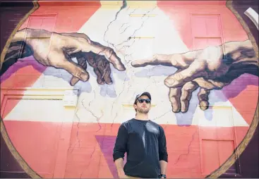  ?? KASSI JACKSON/HARTFORD COURANT ?? Willimanti­c artist Ben Keller stands in front of his latest mural, inspired and based on Michelange­lo’s painting of the hands of God and Adam in the Sistine Chapel, earlier this month on the building next to Cafemantic.