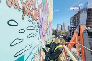  ?? AMY BETH BENNETT PHOTOS/SUN SENTINEL ?? Marcus Borges, a muralist from Funky Paint, checks his drawing Wednesday while he works on a mural designed by Leah Brown on the side of the Extra Space Storage building in the FAT Village neighborho­od of Fort Lauderdale. Borges has been painting the mural for three weeks and says he believes it to be one of the largest in Fort Lauderdale. A man crosses the street as Borges works on the mural behind him.