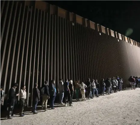  ?? AP ?? People queue against a border wall near Yuma, Arizona, as they wait to apply for asylum after crossing from Mexico