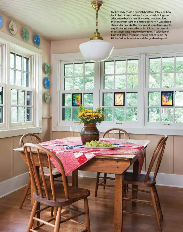  ??  ?? Pat Kennedy chose a mismatched farm table and bowback chairs to set the tone for the casual dining area adjacent to her kitchen. Uncovered windows flood the space with light and natural scenery. A weathered stepladder hosts butter crocks and, sometimes, plants. A quilt draped across the table picks up the colors in the stained-glass window decoration. A selection of blue and green children’s feeding dishes frame the kitchen’s double window and the gardens beyond.