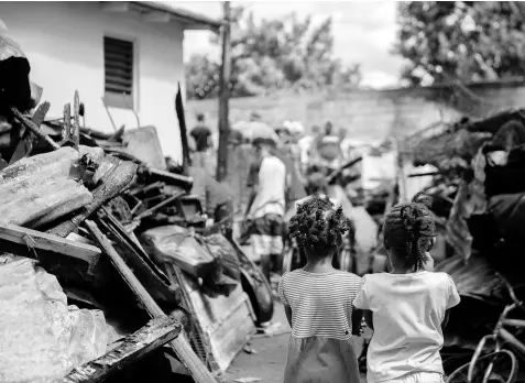  ?? PHOTOS BY GLADSTONE TAYLOR/MULTIMEDIA PHOTO EDITOR ?? Two young girls look on as members of the James Town community in Kencot, St Andrew, clear debris from the fire-gutted area where 15 homes once stood. More than 30 people were left homeless.