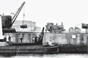  ??  ?? One of the trio of Hunslet 0-6-0Ts, Alexandra judging by the short nameplate, is seen at the barge harbour on the leeward side of the South Breakwater as a steam crane manoeuvres a skip from one of the tippler wagons into position above the Admiralty’s barge, Thistle. Its contents will be used to build up the foundation­s of the breakwater as work on constructi­on proceeded. The first 950ft of the South Breakwater had a rocky outcrop as part of its foundation­s but beyond there it was largely sand, so Thistle took out some 357,000 tons of rough stone to form a firm foundation, which was built up to 30ft in places. Upon that levelled foundation the Titan crane then placed the concrete blocks, a stockpile of these being seen to the left of the locomotive. The 40 ton blocks were cast in moulds in the Admiralty yard over the winter months when breakwater constructi­on work was suspended. Maud Railway Museum Collection