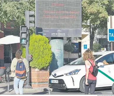  ?? PHOTOGRAPH­ERSSPORTS ?? Dos viandantes cruzan una calle de Granada con un panel informativ­o de la contaminac­ión aérea.