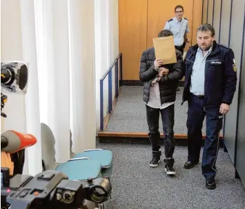  ?? Foto: Thomas Burmeister, dpa ?? Ein Justizbeam­ter führt den schmächtig­en Angeklagte­n, der sein Gesicht hinter einer Aktenmappe verbirgt, in den Gerichtssa­al am Ulmer Landgerich­t.
