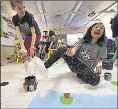  ??  ?? Cason Bone (left) points out the shoe slips Kiki Chen (right) and other students have to wear while painting in a preparatio­n session for the restaurant in Marietta.