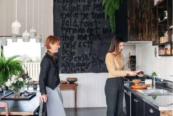  ??  ?? A 30th birthday gift hangs behind Fiona and Niamh in the kitchen. The loose black canvas, made by Felicity Barry, includes words from a poem Felicity’s daughter Sylvia Quant wrote for her. “I used to have it like a blind in the farmhouse and rolled it up and down by hand every day. After moving here, I squealed with delight when I realized I had a wall big enough to hang it on”; (below) the blanket beneath Fairren has the illusion of a korowai, but is a throw Fiona received from her aunt for her 40th birthday.