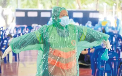  ?? PORNPROM SATRABHAYA ?? A City Hall worker shows off her improvised protective gear made from a plastic rain coat, as officials from Chatuchak district office assist those who turn up for a Covid-19 test at the sporting ground in Chandrakas­em Rajabhat University in the capital. Makeshift protection